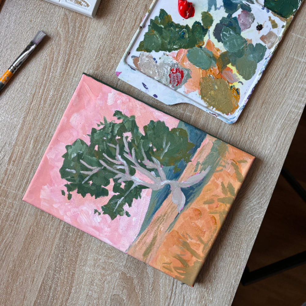 Painting of trees on a pink background with a paint palette and brushes on a wooden surface.